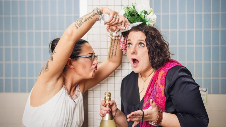 Zwei Frauen in einem Badezimmer, eine sprüht Sekt und die andere schaut überrascht. Die Stimmung ist fröhlich und festlich.