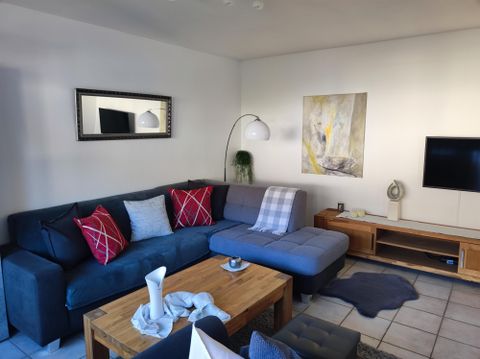 Ein gemütliches Wohnzimmer mit einer dunkelblauen Couch und mehreren Kissen. An der Wand hängt ein modernes Gemälde, und es gibt einen Fernseher und eine Lampe im Raum.