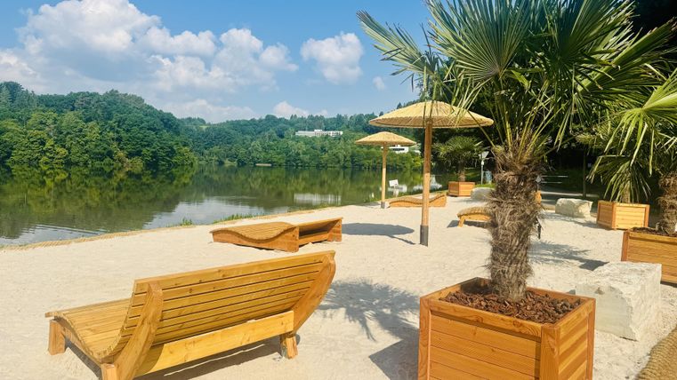 Ein schöner Sandstrand mit Liegen und Palmen. Im Hintergrund ist ein ruhiger See und grüne Bäume sichtbar.