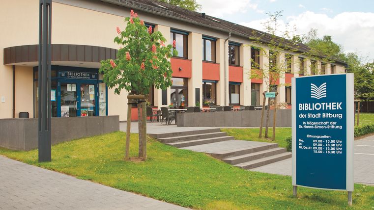 Eine moderne Bibliothek mit einem großen Schild und grünen Pflanzen. Die Fassade ist freundlich gestaltet und bietet einen einladenden Zugang.
