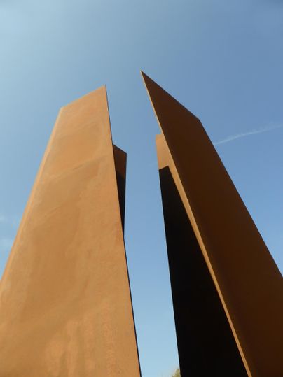 Zwei hohe, spitze Stahlstelen vor einem blauen Himmel. Die Stelen sind rostfarben und stehen auseinander.
