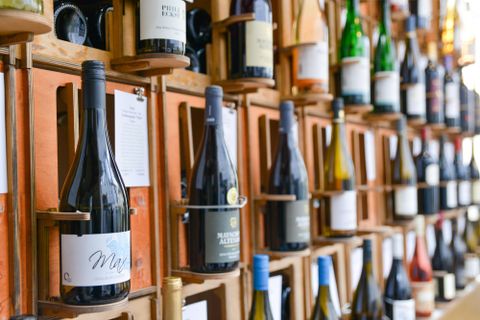 Een wijnlokaal met een muur vol wijnflessen. De omgeving is helder en vriendelijk, met een plant in de hoek.