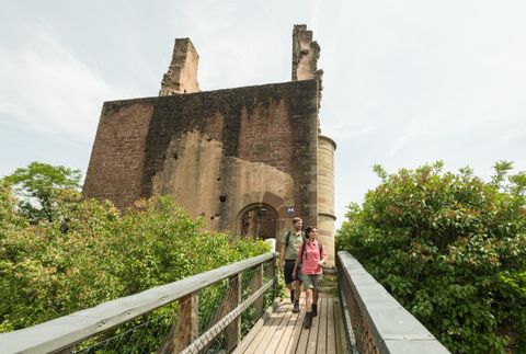 Une vieille ruine de château entourée d'arbres. Deux randonneurs marchent sur une passerelle en bois en direction de la ruine.