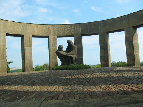 Eine monumentale Skulptur von zwei Figuren, die sich umarmen, steht im Mittelpunkt eines runden Steingebäudes. Der Himmel ist blau mit wenigen Wolken, und die Umgebung ist grün und friedlich.