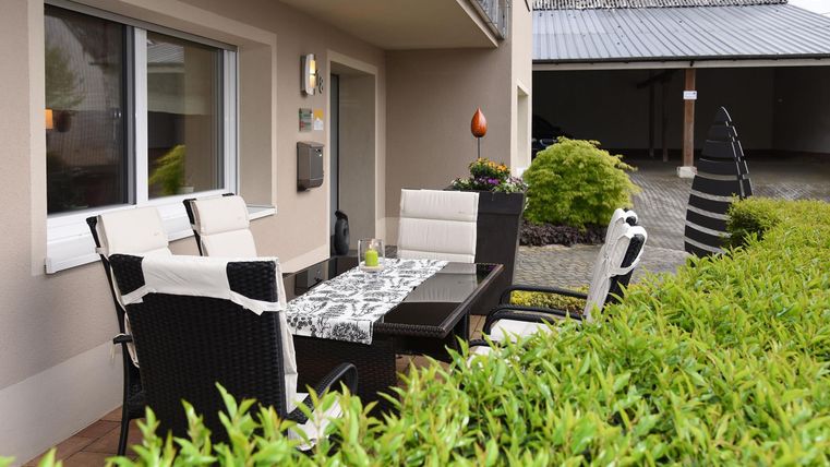 Un espace extérieur confortable avec une zone de repas en rotin noir et une table. Une lumière verte et des plantes entourent l'espace, idéal pour les repas en plein air.