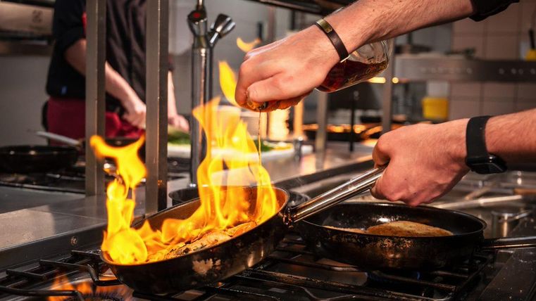 Ein Koch in einer Küche bereitet ein Gericht zu. Flammen leuchten aus einer Pfanne, während er eine Flüssigkeit hinzufügt.