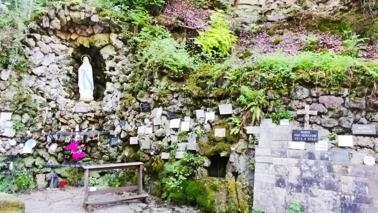 Eine ruhige Grotte mit einer Statue und vielen Erinnerungsplaketten. Grüne Pflanzen umgeben den Ort, der eine spirituelle Atmosphäre ausstrahlt.