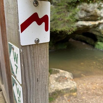 Un panneau indicateur avec un symbole rouge sur un poteau en bois. En arrière-plan, on peut voir une petite étendue d'eau et des rochers.