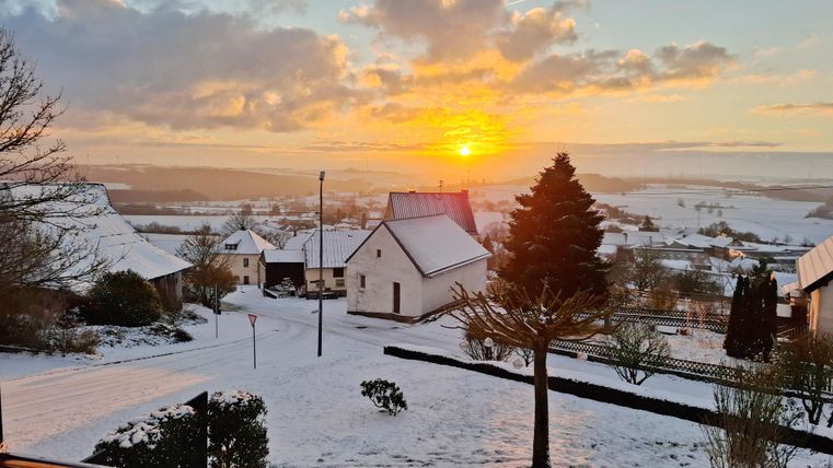 Un paysage enneigé avec des maisons et des arbres. En arrière-plan, le soleil brille à l'horizon.