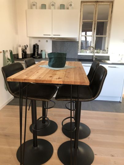 Ein moderner Essbereich mit einem hohen Holztisch und vier schwarzen Barhockern. Im Hintergrund sind Küchenmöbel und eine große Fensterfront sichtbar.