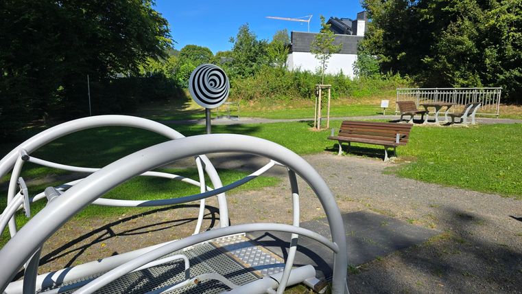 Ein Spielplatz mit modernen Geräten und einer Wiese im Hintergrund. Die Umgebung ist grün und es ist sonnig.