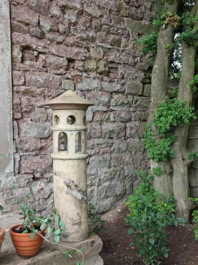 Een oude pozzolan met een decoratieve toren staat tegen een stenen muur. Daarnaast zijn enkele bloempotten en groene planten te zien.