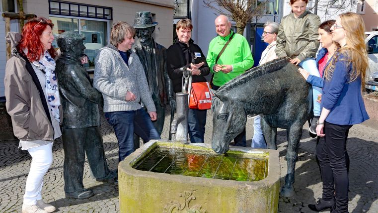 Eine Gruppe von Menschen steht um eine Statue mit einem Pferd und mehreren Figuren. Im Hintergrund sind einige Gebäude und ein Brunnen zu sehen.