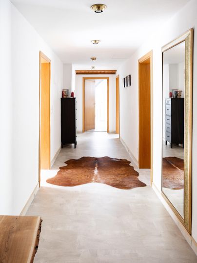 A bright hallway with white walls and a large mirror. A cowhide lies on the floor, and there are wooden and cupboard furniture on the walls.