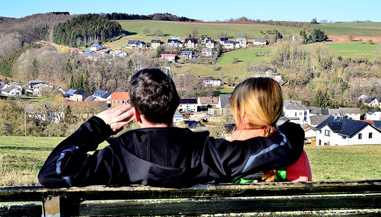 Ein Paar sitzt auf einer Bank und blickt auf das Dorf Baustert in einer hügeligen Landschaft.
