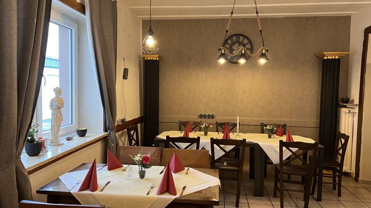 Ein gemütlicher Speiseraum mit dekorierten Tischen und braunen Holzstühlen. An den Wänden hängen stilvolle Vorhänge und eine Uhr sorgt für ein besonderes Ambiente.