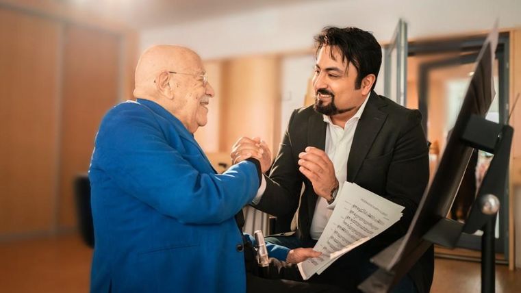 Ein älterer Mann in einem blauen Anzug und ein jüngerer Mann im Anzug lächeln sich an und halten Händchen. Im Hintergrund sind unscharfe Möbel und ein Notenblatt zu sehen.