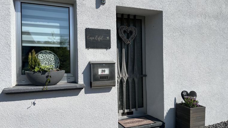 Un hall d'entrée avec un mur blanc et une porte décorative. À côté de la porte se trouve un pot de fleurs et une boîte aux lettres.