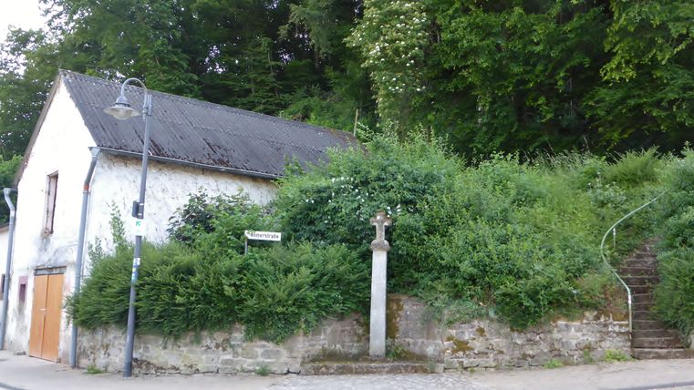 Ein altes Gebäude an einer Straßenecke mit einem Kreuz und Treppen, umgeben von viel Grün.