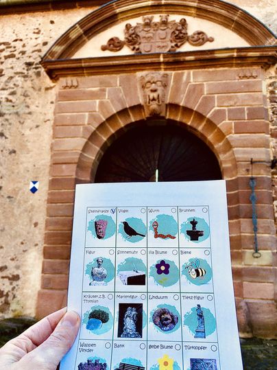 Ein historisches Gebäude mit einer großen, runden Tür und einem Wappen oberhalb. In der Hand hält jemand ein Papier mit verschiedenen Abbildungen dazu.