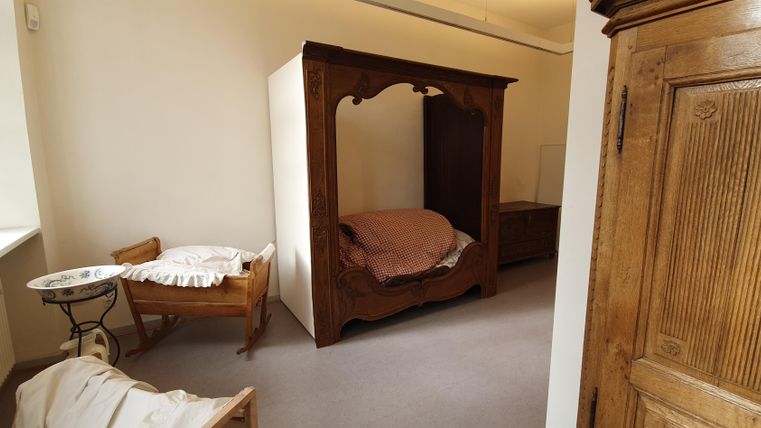 A simple, bright room with a wooden wardrobe and a large bed. Next to the bed, there is a small table and a chair.