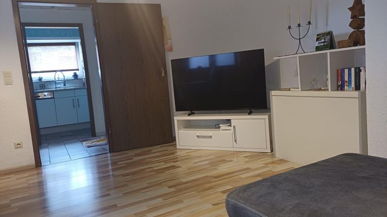 Ein gemütliches Wohnzimmer mit einem Fernseher und einem modernen TV-Regal. Der Raum hat einen hellen Holzboden und führt zu einer Küche.