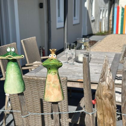Zwei bunte Figuren in Form von Fröschen, die an einem Zaun hängen. Im Hintergrund ist eine gemütliche Terrasse mit Gartenmöbeln zu sehen.