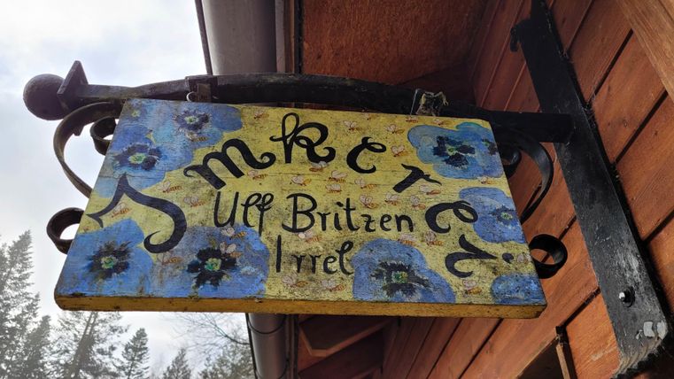 Een handgeschilderd bord met blauwe bloemen en de tekst "Imkerei auf Brütten Irrel". Het hangt aan een houten gebouw en straalt een rustieke charme uit.