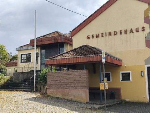 Ein modernes Gemeindehaus mit einem schlichten Design und einem gepflasterten Vorplatz. Die Fassade ist in warmen Farben gehalten und bietet einen einladenden Eindruck.
