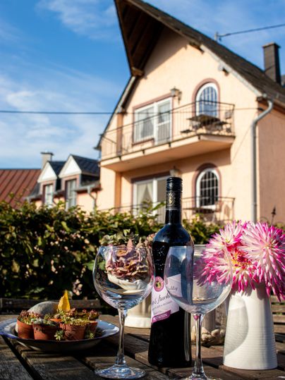 Ein schöner Tisch im Freien mit einer Flasche Wein, Gläsern und einer bunten Blumenvase. Im Hintergrund steht ein charmantes Haus unter klarem Himmel.