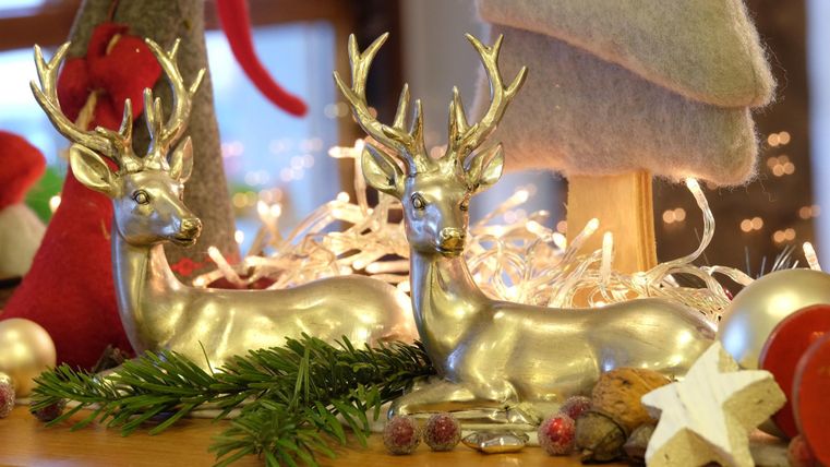 Zwei silberne Rentierfiguren stehen auf einem dekorierten Tisch. Um sie herum sind Weihnachtsdekorationen wie Zweige, Sterne und Glanzkugeln.
