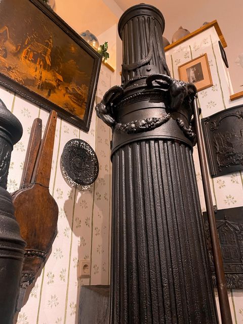 A historic black stove in a room with decorative elements and pictures on the wall. The stove is tall and has an ornate structure.