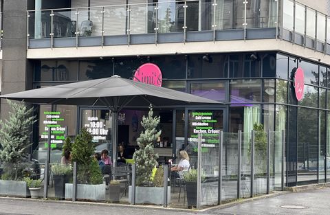 Un café moderne avec de grandes fenêtres et une terrasse. Il y a des places assises à l'extérieur et des plantes décoratives.