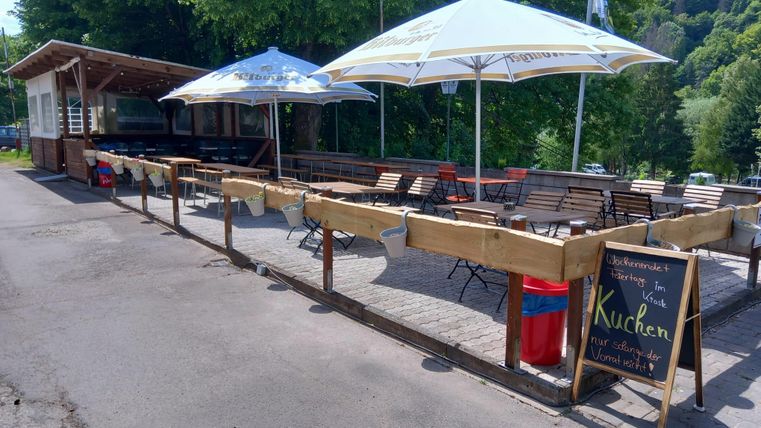 Een gezellig café met houten stoelen en tafels buiten. Paraplu's beschermen de gasten tegen de zon, en een bord wijst op taarten.