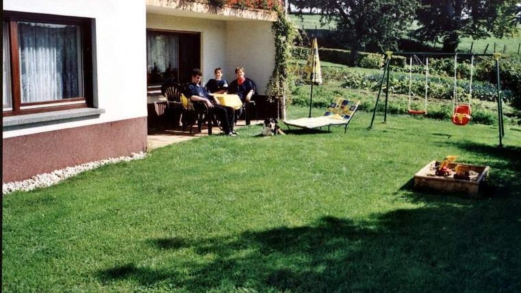 Une pelouse verte derrière une maison avec une terrasse. Trois personnes sont assises à la table et se détendent, tandis qu'en arrière-plan, il y a une balançoire et un petit jardin.