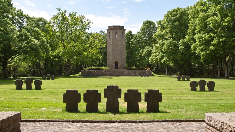 Kriegsdenkmal mit Steinkreuzen und Turm im Hintergrund, umgeben von Bäumen.