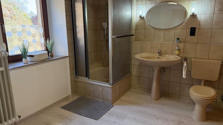 Ein modernes Badezimmer mit einer Dusche, einem Waschbecken und einer Toilette. Die Wände sind gefliest und es gibt ein Fenster mit Pflanzen.