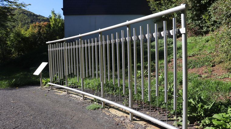 Ein gebogener Metallzaun steht an einem Weg. Im Hintergrund sind Bäume und ein Gebäude sichtbar.