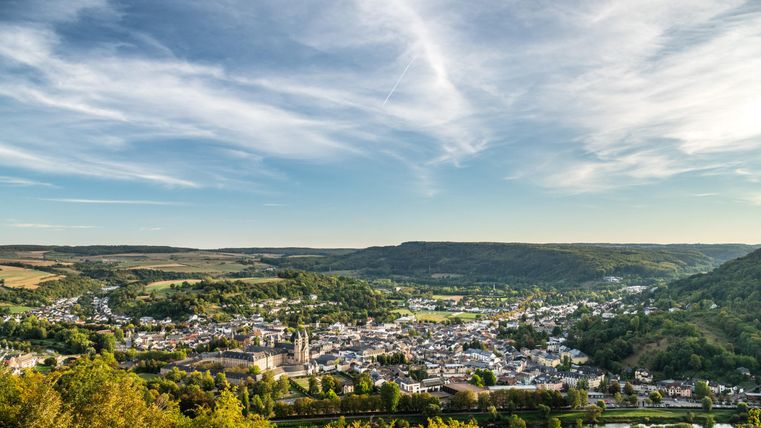 Eine malerische Stadtlandschaft umgeben von grünen Hügeln und Wäldern. Der Himmel ist klar mit leichten Wolken und vermittelt eine friedliche Atmosphäre.