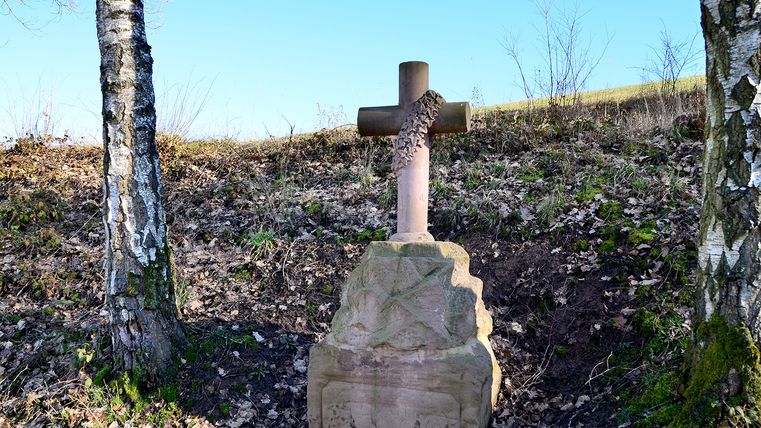Steinernes Wegekreuz mit Efeu bewachsen zwischen zwei Bäumen.