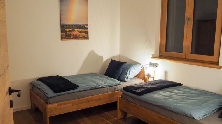 Ein einfaches Schlafzimmer mit zwei Einzelbetten aus Holz. An der Wand hängt ein Bild mit einem Regenbogen und es gibt ein Fenster mit natürlichem Licht.