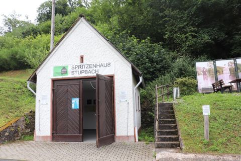 Ein kleines, weißes Gebäude mit offener Tür, das als "Hirtenhaus Stupbach" gekennzeichnet ist. Es liegt an einem bewaldeten Hang und hat Treppen, die zu einem weiteren Bereich führen.