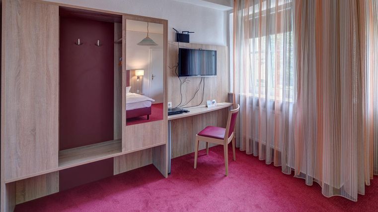 Une chambre d'hôtel simple avec un tapis rouge, un bureau et une télévision. La pièce a de grandes fenêtres avec des rideaux et une armoire.