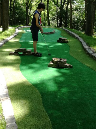 Ein Mensch spielt Minigolf auf einem grünen Platz im Wald. Um ihn herum sind Bäume und verschiedene Hindernisse aus Steinen.