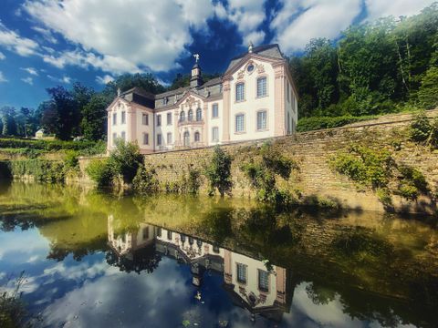 Ein hübsches Schloss am Wasser, umgeben von Bäumen. Die Wolken reflektieren sich im ruhigen Wasser.