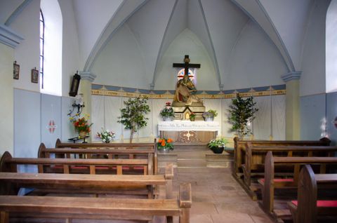 Ein kirchlicher Innenraum mit Holzbänken und einem Altar. Im Hintergrund steht eine Statue von Jesus am Kreuz, umgeben von Blumen und Pflanzen.