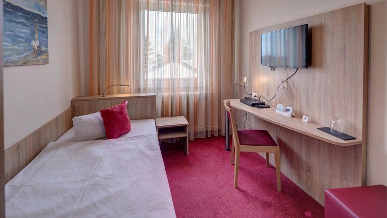 Une chambre simple avec un lit simple et un bureau. De grandes fenêtres laissent entrer beaucoup de lumière et créent une atmosphère lumineuse.