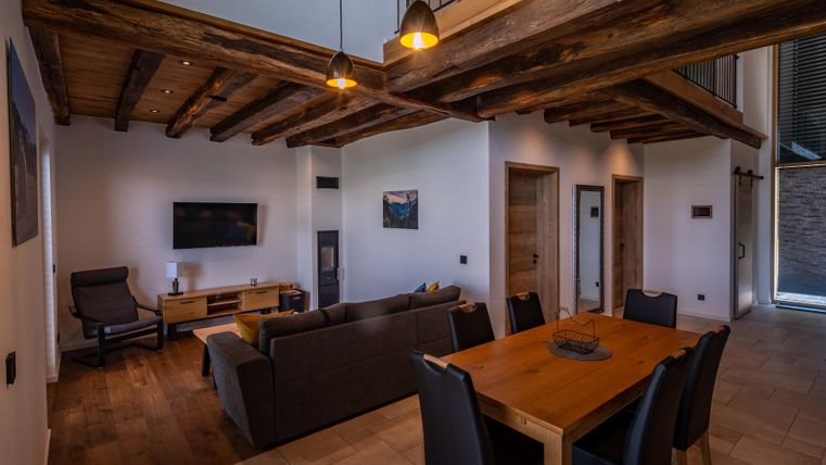 Ein modernes Wohnzimmer mit Holzdecken und einem stilvollen Esstisch. Der Raum ist hell und einladend, mit gemütlichen Möbeln und einer großen Fensterfront.