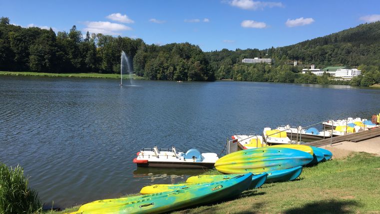 Un lac calme avec des kayaks colorés sur la rive. À l'arrière-plan, on peut voir des arbres et un ciel bleu.