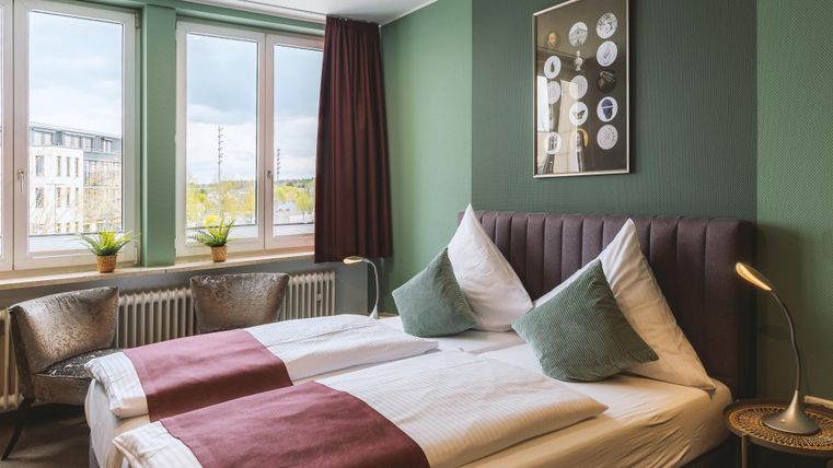 Ein modernes Schlafzimmer mit zwei Betten, dekoriert in sanften Farben. Große Fenster sorgen für viel Tageslicht und eine angenehme Atmosphäre.